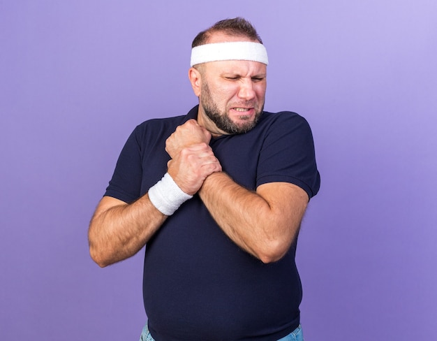 Pijnlijke volwassen slavische sportieve man met hoofdband en polsbandjes met hand geïsoleerd op paarse muur met kopieerruimte