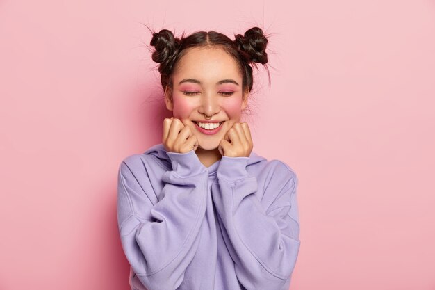 Pinup Japanse vrouw met twee gekamde haarbroodjes raakt wangen, heeft een gladde huid, draagt levendige roze make-up, piercing in de neus, draagt een sweatshirt, glimlacht positief, geïsoleerd op roze achtergrond.