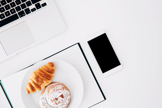 Plaat van croissant en broodjes op agenda met smartphone en laptop op witte achtergrond