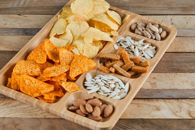 Gratis foto plaat van diverse snacks op een houten tafel. chips, crackers, amandelen, pistachenoten, zonnebloempitten.