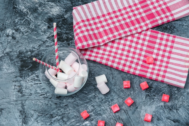 Plat lag marshmallows en suikerriet in pot met snoepjes en rood pastel tafelkleed op donkerblauw marmeren oppervlak. horizontaal