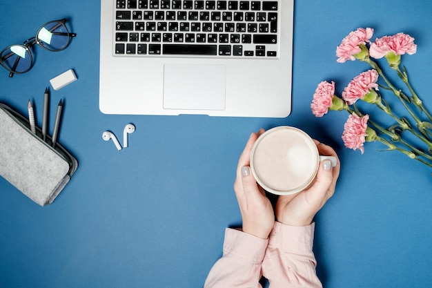 Gratis foto platliggend vrouwenbureau vrouwenhanden met koffie en laptop roze anjer briefpapier op blauwe tafel bovenaanzicht