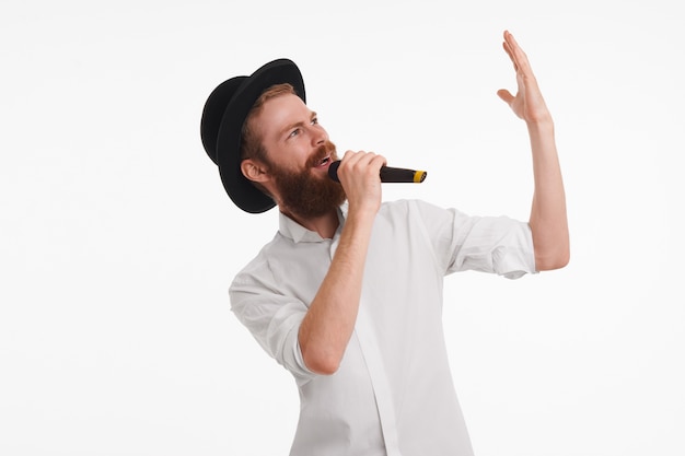 Popzanger met wazige baard die emotioneel gebaart tijdens het optreden met een microfoon. aantrekkelijke bebaarde jonge mannelijke entertainer met zwarte hoed en een wit overhemd met microfoon, iets aankondigend