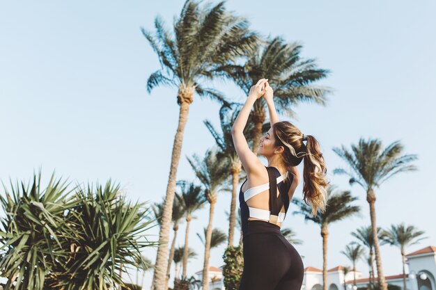 Portret aantrekkelijke jonge sportvrouw die zich uitstrekt over palmbomen, blauwe hemel. Zonnige ochtend, training, motivatie, fitness, training, ontspanning.