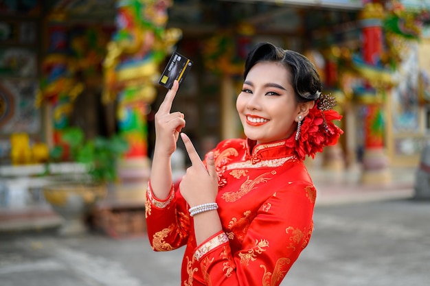 Portret aziatische mooie vrouw die een cheongsam draagt die lacht en poseert, toont creditcard bij heiligdom op chinees nieuwjaar