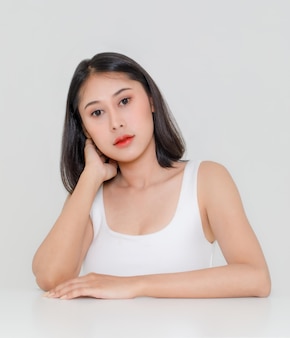 Portret close-up studio shot van millennial aziatisch kort zwart haar model met make-up rode lip in tank top hemd zittend aan tafel poseren hand in hand op wang kijken camera op witte achtergrond.