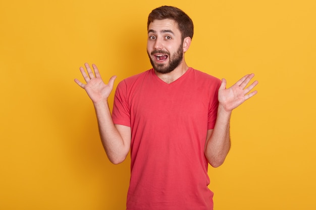 Portret dat van de mens wordt verrast, knap mannetje dat zijn handen uitspreidt, geïsoleerd stellen over gele muur, aantrekkelijke ongeschoren kerel die rode toevallige t-shirt draagt. het concept van menselijke emoties.