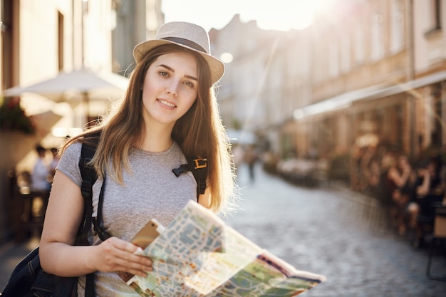 Portret dat van een vrouw de wereld reist met behulp van een kaart en een tablet, die zich in een kleine Europese stad bevindt die camera bekijkt.