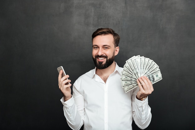 Portret die van de vrolijke mens in wit overhemd veel de munt van de gelddollar winnen die zijn smartphone gebruiken, blij over donkergrijs zijn
