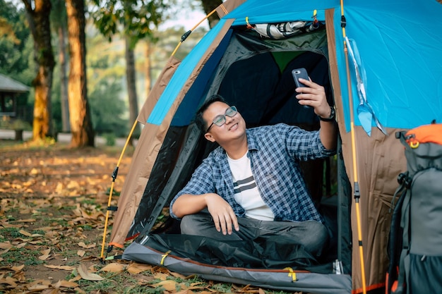 Portret gelukkige Aziatische man bril maken van een video-oproep met smartphone in tent camping Koken set voorgrond Outdoor koken reizen camping lifestyle concept