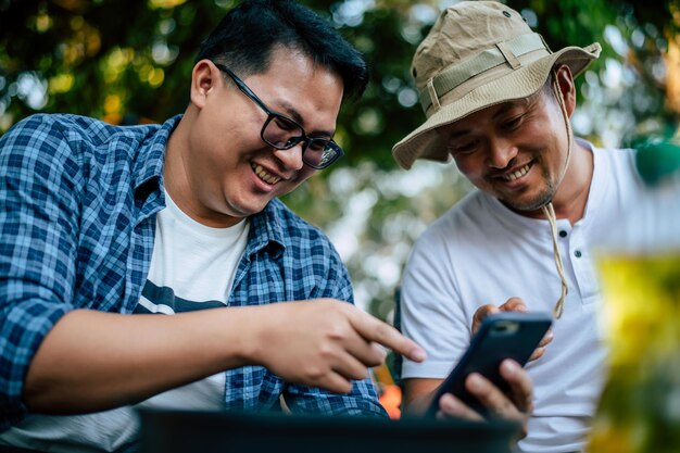 Portret gelukkige Aziatische man vrienden zittend op een stoel in het kamp met praten en kijken naar smartphone Koken set voorgrond Buiten koken reizen camping lifestyle concept