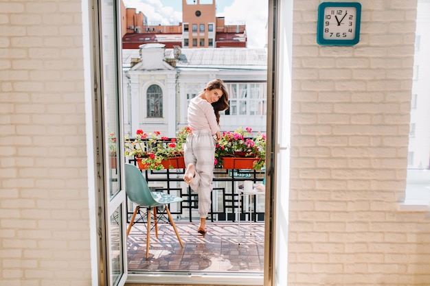 Portret jong meisje in pyjama verplaatsen op balkon in stad in zonnige ochtend. Haar lange haren vliegen op de wind, ze glimlachte.