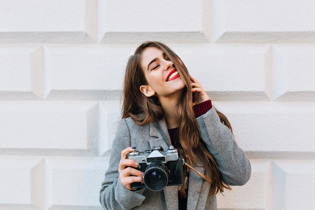 Portret jong meisje met lang haar en rode lippen in grijze vacht op muur. Ze houdt de camera vast, houdt de ogen gesloten en lacht.