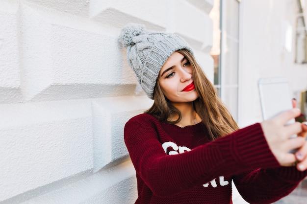 Gratis foto portret jong meisje met lang haar selfie maken op grijze muur. ze draagt een gebreide muts en heeft rode lippen.