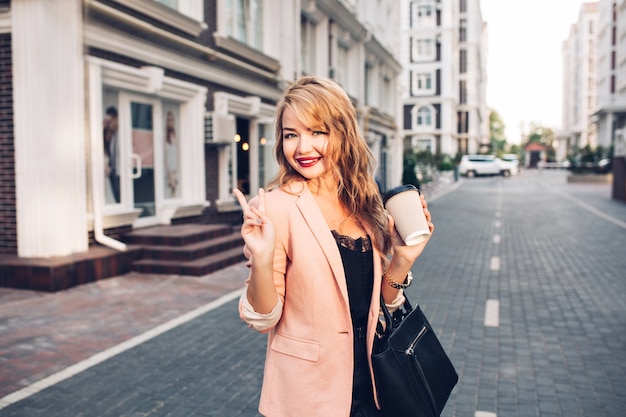 Portret modieuze blonde vrouw met lang haar wandelen in koraal jasje op straat. Ze houdt een kopje koffie vast