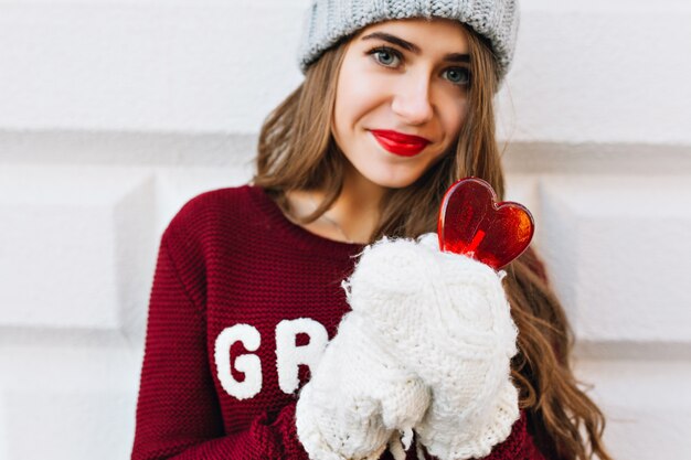 Portret mooi meisje in marsala sweater en witte handschoenen op grijze muur. Ze draagt een gebreide muts, houdt een hartlolly vast en kijkt.