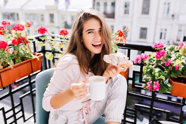 Portret mooi meisje met lang haar ontbijten op balkon in de ochtend in de stad. ze houdt een kopje, croissant, lachend vast.