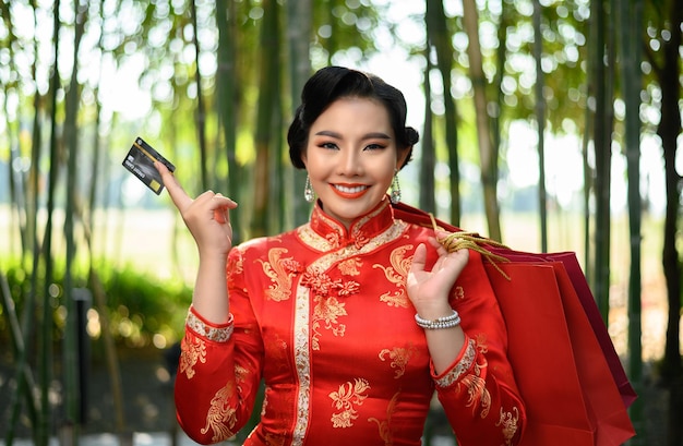 Gratis foto portret mooie aziatische vrouw in een chinese cheongsam met boodschappentassen en creditcard in de hand met geluk in bamboebos