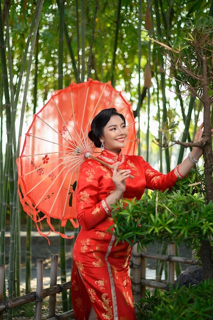 Portret mooie aziatische vrouw in een chinese cheongsam poseren met mooie rode papieren paraplu op bamboebos, kopieer ruimte