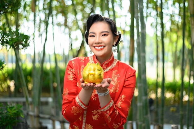 Portret mooie aziatische vrouw in een chinese cheongsam poseren met spaarvarken op bamboebos