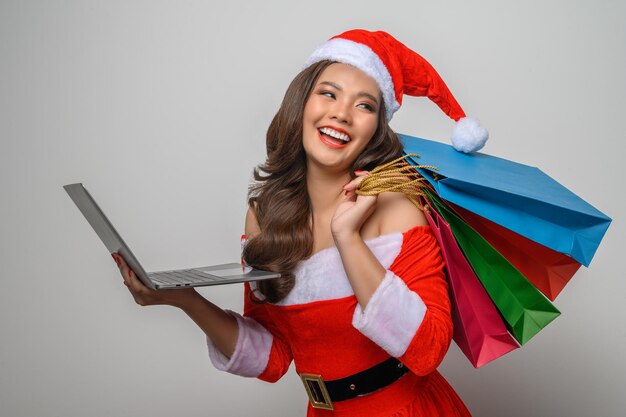 Portret mooie aziatische vrouw in rode kerstman kostuum poseren met laptopcomputer en boodschappentassen in de hand