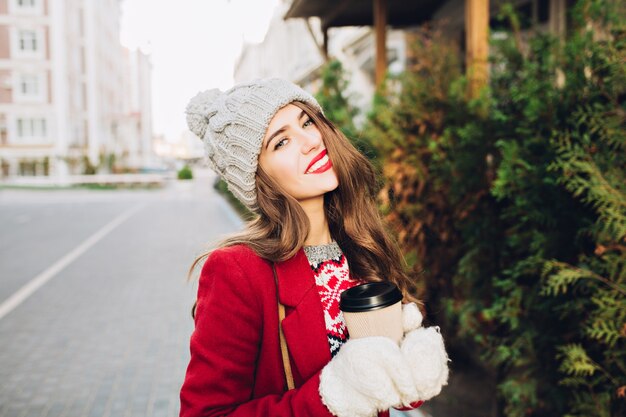 Portret mooie brunette meisje met lang haar in een rode jas lopen op straat in de stad. Ze houdt koffie voor onderweg vast in witte handschoenen, glimlachend met rode lippen.
