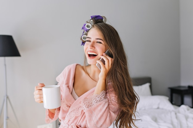 Portret mooie huisvrouw in roze badjas met krultang thuis. Ze sprak aan de telefoon en lachte.