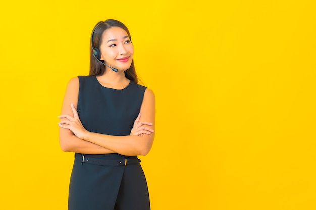 Portret mooie jonge Aziatische bedrijfsvrouw met de klantenzorg van het hoofdtelefooncall centre op gele achtergrond