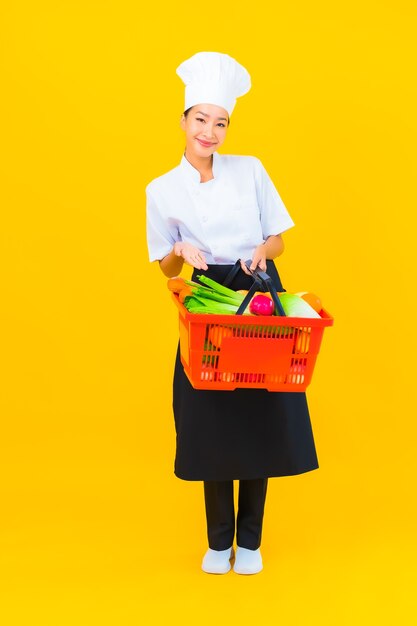 Portret mooie jonge aziatische chef-kokvrouw met kruidenierswinkelmand van supermarkt op gele geïsoleerde background