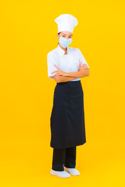 Portret mooie jonge aziatische vrouw chef-kok draagt een masker ter bescherming van covid19 of coronavirus op gele geïsoleerde achtergrond