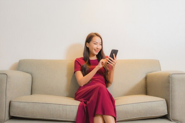 Portret mooie jonge Aziatische vrouw die slimme mobiele telefoon op bank in woonkamerbinnenland met behulp van