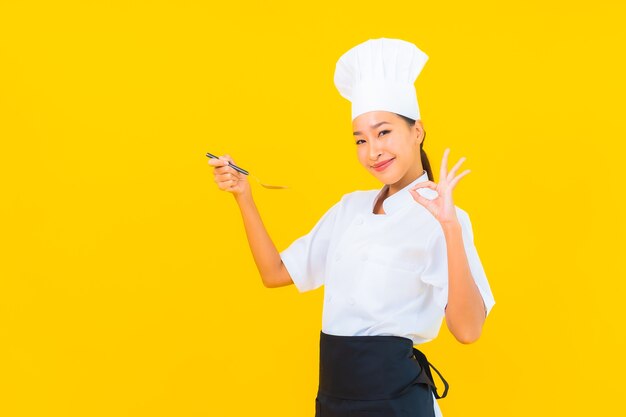 Portret mooie jonge aziatische vrouw draagt chef-kok of kookuniform met lepel en vork op gele geïsoleerde achtergrond