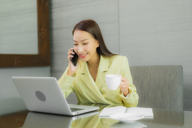 Portret mooie jonge Aziatische vrouw gebruik computer laptop met slimme mobiele telefoon op werktafel op interieur kamer