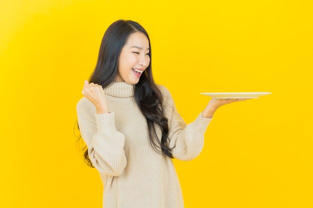 Portret mooie jonge aziatische vrouw glimlacht met lege bordschotel op gele muur