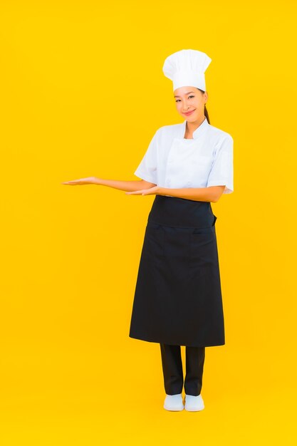 Portret mooie jonge aziatische vrouw in chef-kok of kok uniform met hoed op gele geïsoleerde achtergrond