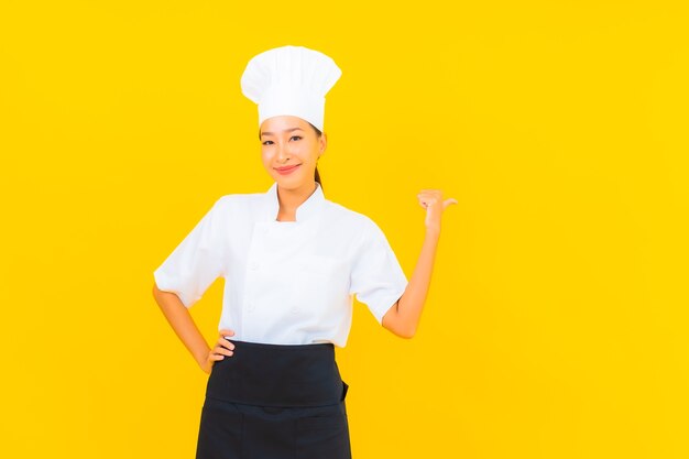 Portret mooie jonge Aziatische vrouw in chef-kok of kok uniform met hoed op gele geïsoleerde achtergrond