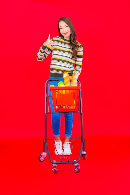 Gratis foto portret mooie jonge aziatische vrouw met kruidenierswinkelmandje van supermarkt op rode geïsoleerde muur