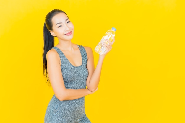 Portret mooie jonge Aziatische vrouw met sportkleding en flessenwater op gele muur