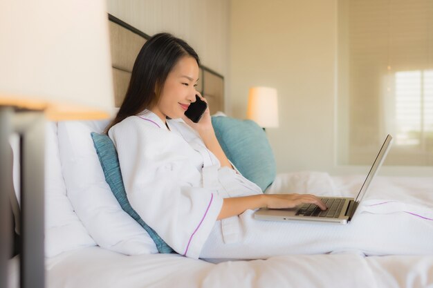 Portret mooie jonge Aziatische vrouwen die laptop of computer met mobiel op bed met behulp van