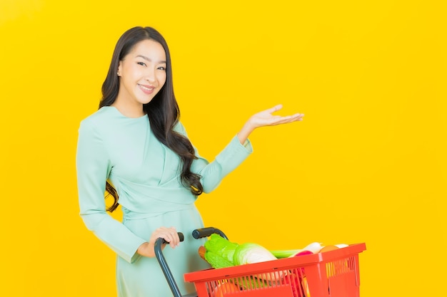 Portret mooie jonge aziatische vrouwenglimlach met kruidenierswinkelmand van supermarkt op geel