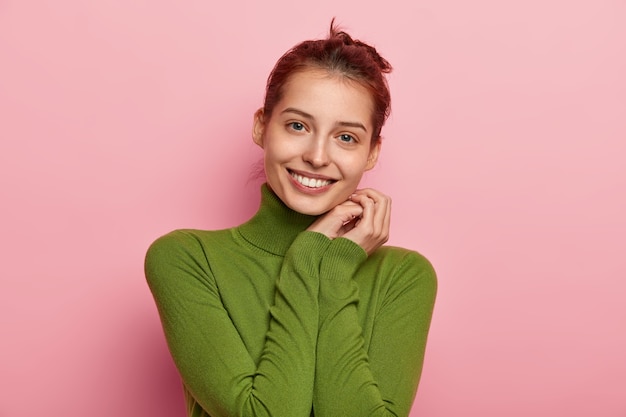 Portret van aangenaam uitziende jonge vrouw met brede glimlach, houdt handen in de buurt van wang, gekleed in casual poloneck, kijkt vrolijk, geniet van goed nieuws, vormt tegen roze achtergrond.