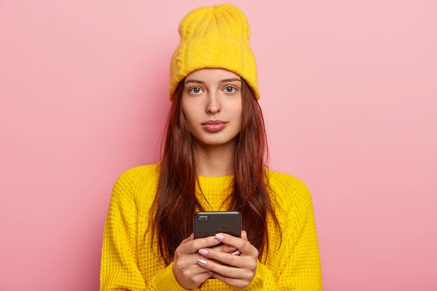 Portret van aangenaam uitziende vrouw kijkt serieus, gebruikt moderne mobiele telefoon, draagt gele hoed en winter trui, vormt op roze achtergrond, draagt geen make-up