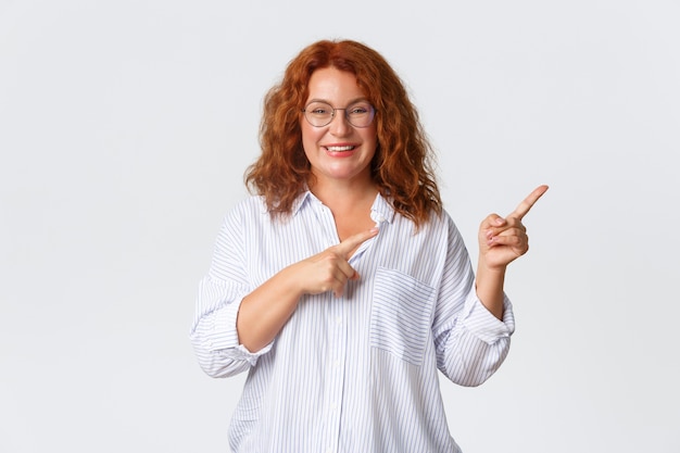 Portret van aangename glimlachende vrouw van middelbare leeftijd met rood haar, bril en blouse met reclame, klant van bedrijf beveelt product of dienst aan, naar rechts wijzend.