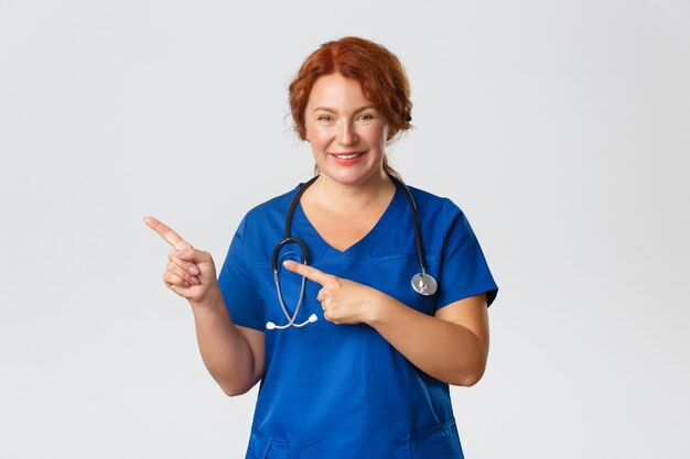 Portret van aangename vrouwelijke arts, roodharige verpleegster in blauwe scrubs die vrolijk kijkt, kliniekbanner, ziekenhuisadvertentie, wijzende vinger naar links toont