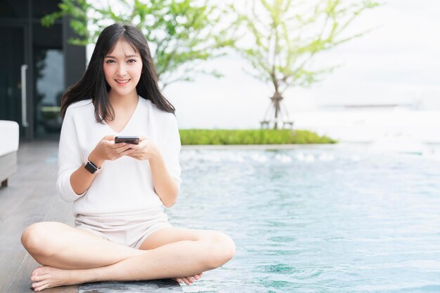 Portret van aantrekkelijke Aziatische lang zwart haar casual jurk glimlach hand gebruik smartphone zitten ontspannen in de buurt van zwembad tuin met het bouwen van stad achtergrond communicatie en technologie ideeën concept
