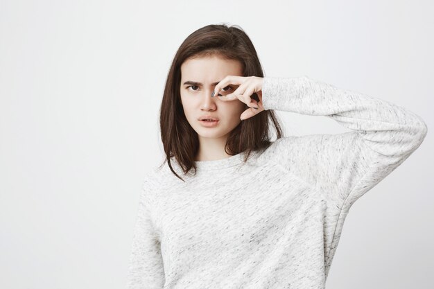 Portret van aantrekkelijke ernstig ogende vrouw in witte sweater die gebaar dichtbij oog met geconcentreerde en zekere uitdrukking maakt.