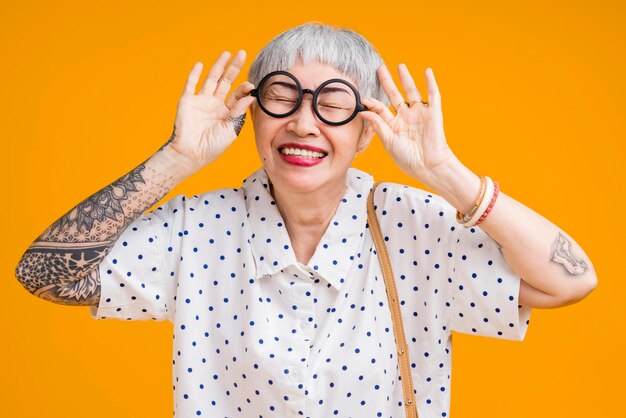 Portret van aantrekkelijke funky Aziatische tatoeage vrolijke grijsharige oudere oude senior vrouw aanraken bril geïsoleerd over gele pastel achtergrond kleurenverrassing en over verlaten gevoel expressie