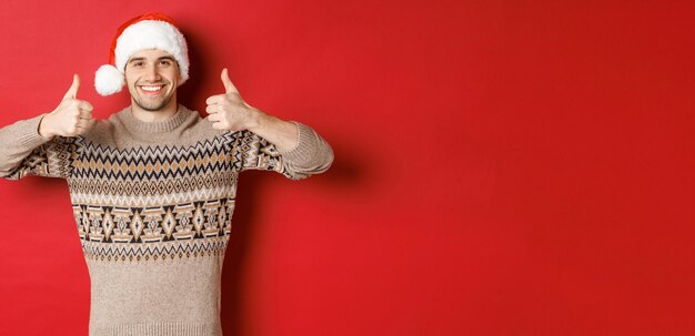 Portret van aantrekkelijke gelukkige man in kerstmuts en trui die thumbsup in goedkeuring toont en glimlachend vrolijk kerstfeest wenst over rode achtergrond