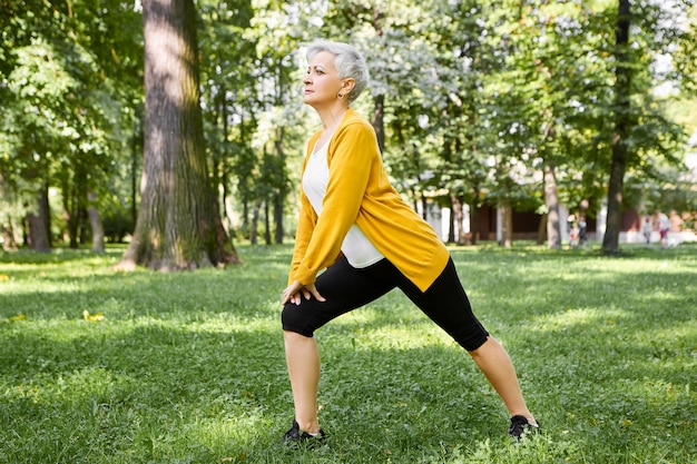 Portret van aantrekkelijke gezonde zestig jaar oude vrouw staande op een been en die zich uitstrekt in pilates pose. Grijze haren senior vrouw in sportkleding kant lunges doen op gras in stadspark op zonnige dag