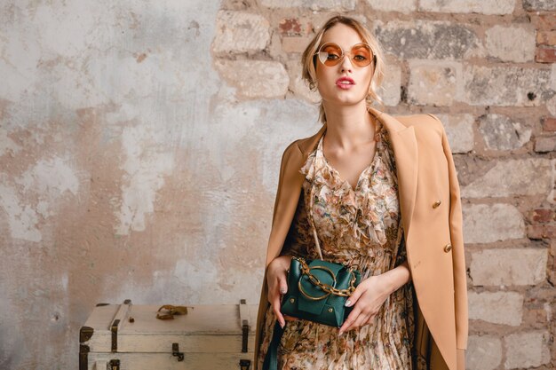 Portret van aantrekkelijke glimlachende stijlvolle blonde vrouw in beige jas wandelen in straat tegen vintage muur